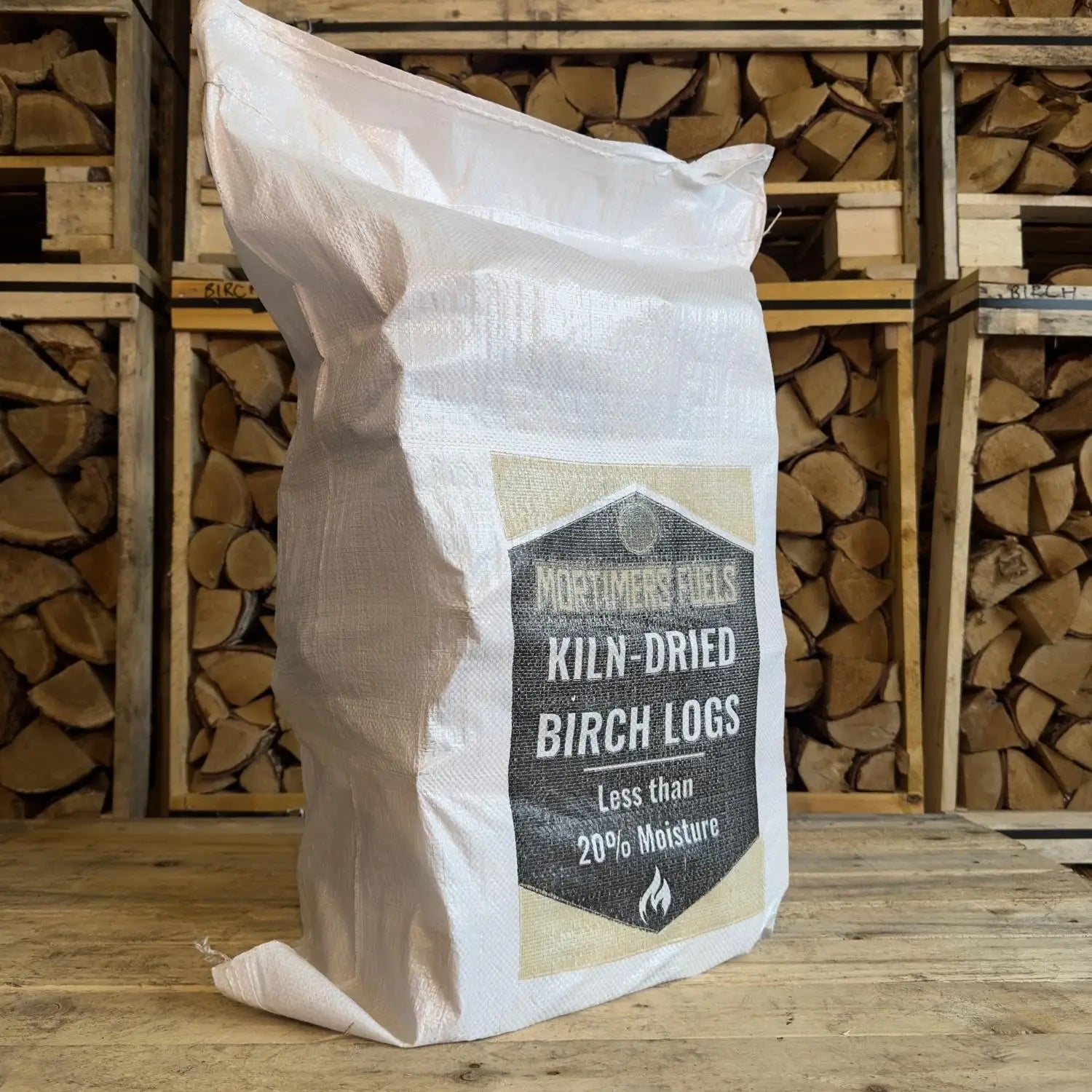 Bag of kiln-dried birch logs with a visible brand label against a background of stacked logs.