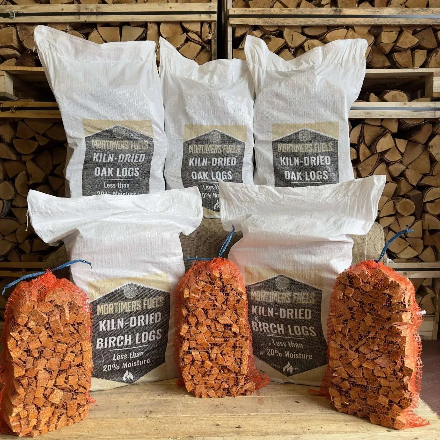 Bags of Mortimer's Fuels kiln-dried oak and birch logs with wooden logs displayed in front.