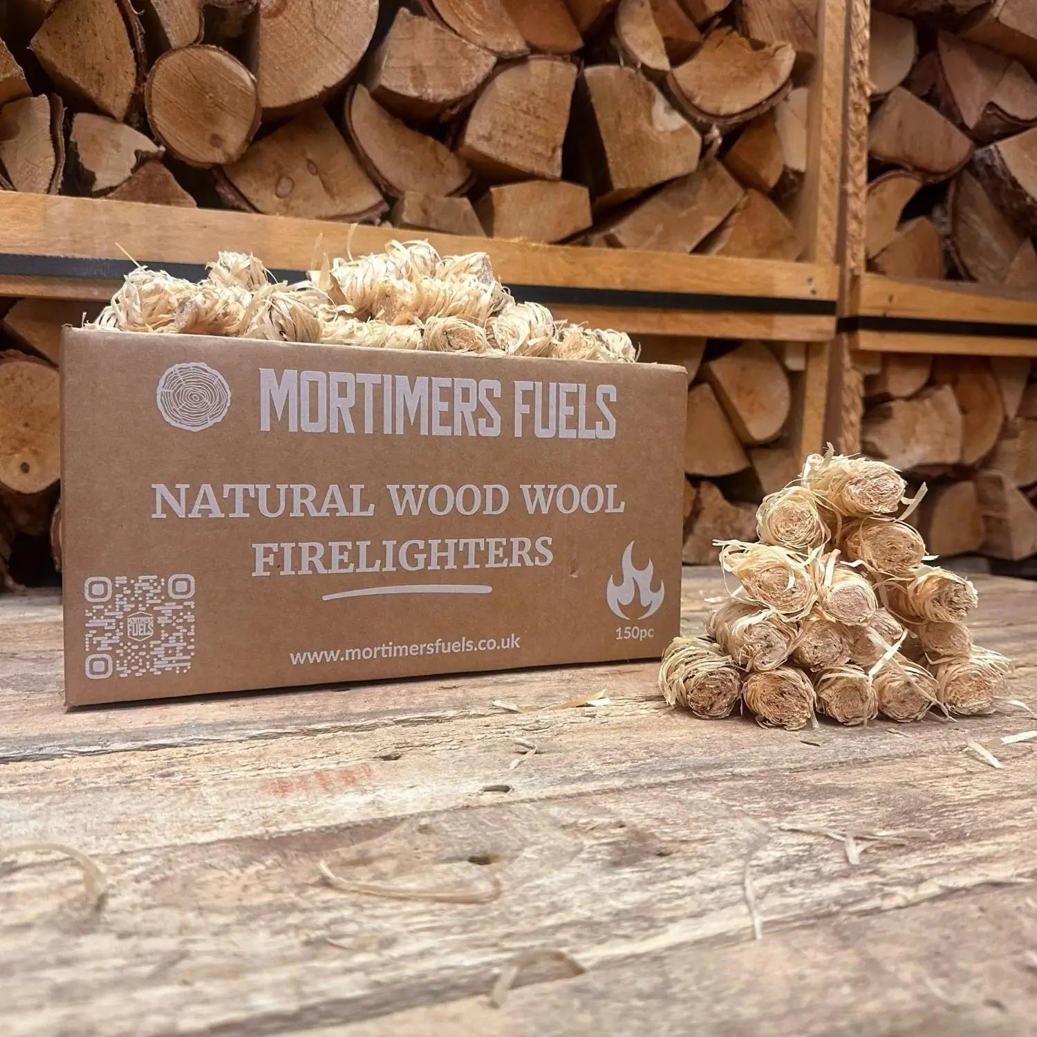 Box of Mortimer's Fuels natural wood wool firelighters with firelighters on a wooden surface and stacked logs in the background.
